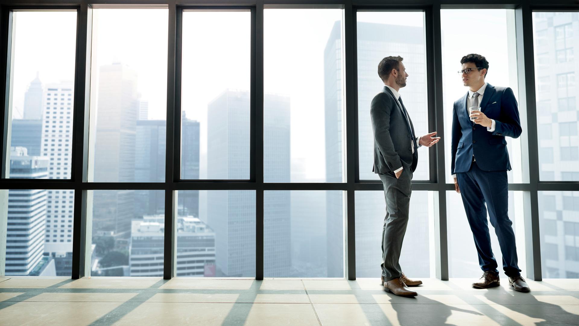 Two businessmen in suits talking by large office windows overlooking a city skyline.